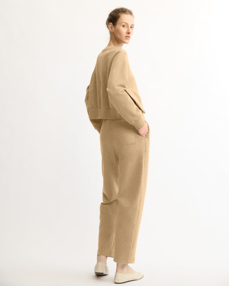 A person with light hair tied back stands sideways with hands in pockets, wearing Arlo Mott Baltic Barrel Leg Sweatpants in Khaki and matching jacket, glancing over their shoulder in slip-on shoes against a plain backdrop.