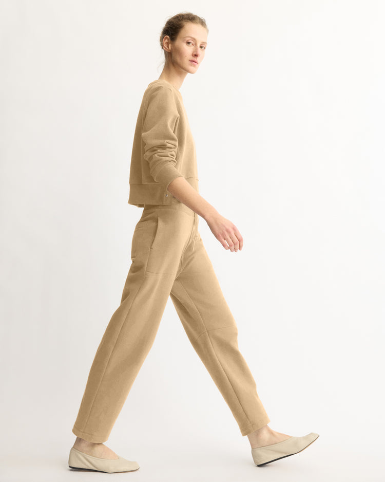 A woman with light hair in a bun walks sideways against a plain white background, wearing Arlo Mott’s Baltic Barrel Leg Sweatpants in Khaki, paired with a beige long-sleeve top and light beige flats.