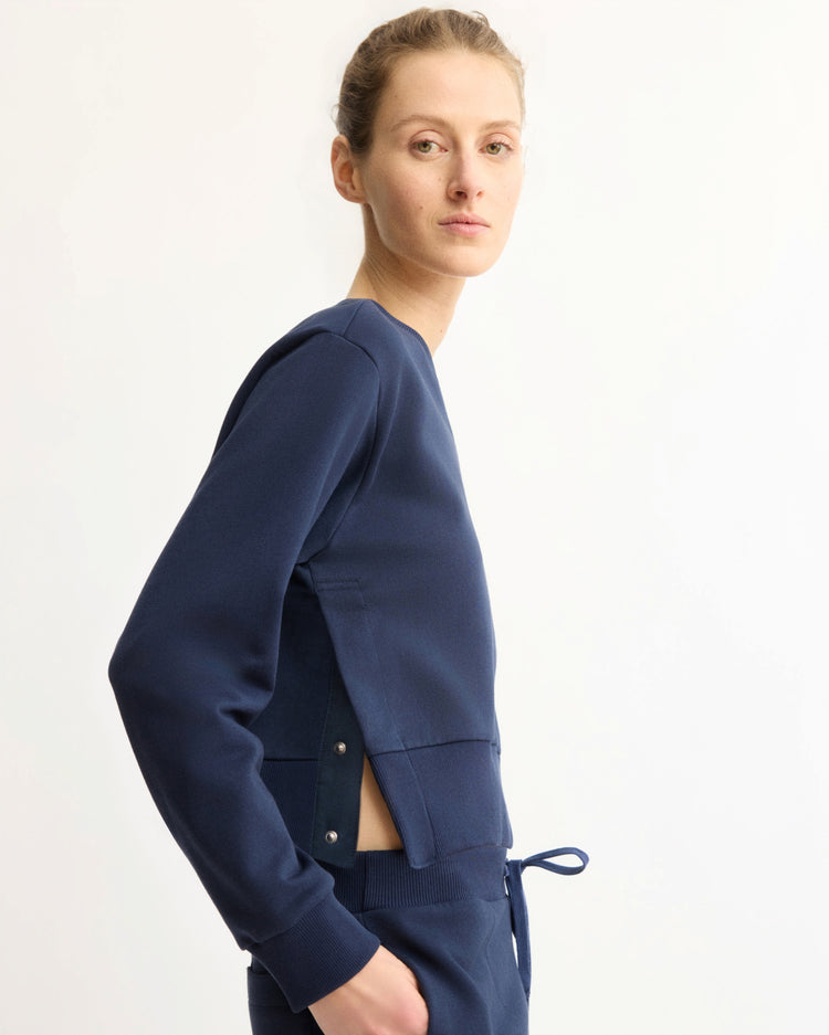 A person wearing the Arlo Mott Kent Crewneck With Snaps in Navy stands sideways against a plain light background, looking at the camera. The crewneck features side snap detailing and a waist tie.