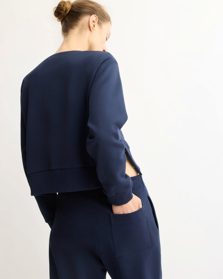 A person with light skin and brown hair in a bun wears the Arlo Mott Kent Crewneck With Snaps in Navy, standing with their back slightly turned toward the camera against a plain light background.