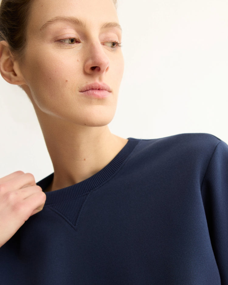 A person with light skin and tied-back hair looks to the side while wearing the Arlo Mott Kent Crewneck With Snaps in Navy against a plain, light background.