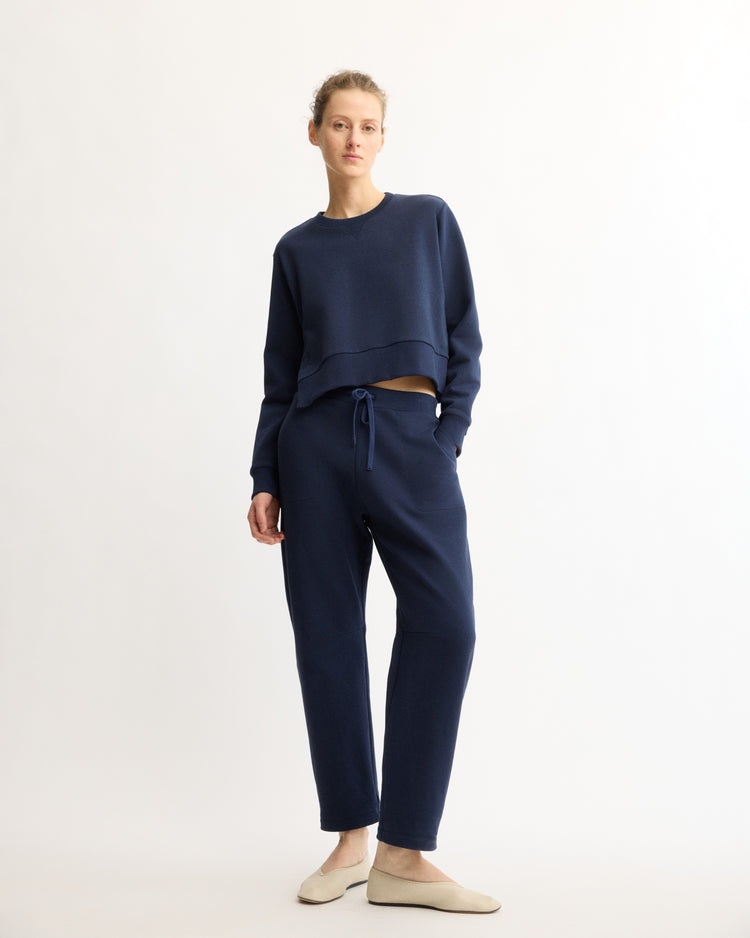 A woman stands against a light background, wearing Arlo Mott Baltic Barrel Leg Sweatpants in Navy with a loose-fitting navy long-sleeve top and beige slip-on shoes.
