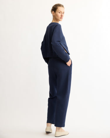 A woman stands sideways against a white background, looking over her shoulder. She wears Arlo Mott's Baltic Barrel Leg Sweatpants in Navy with a matching navy Italian terry top and light slip-on shoes.