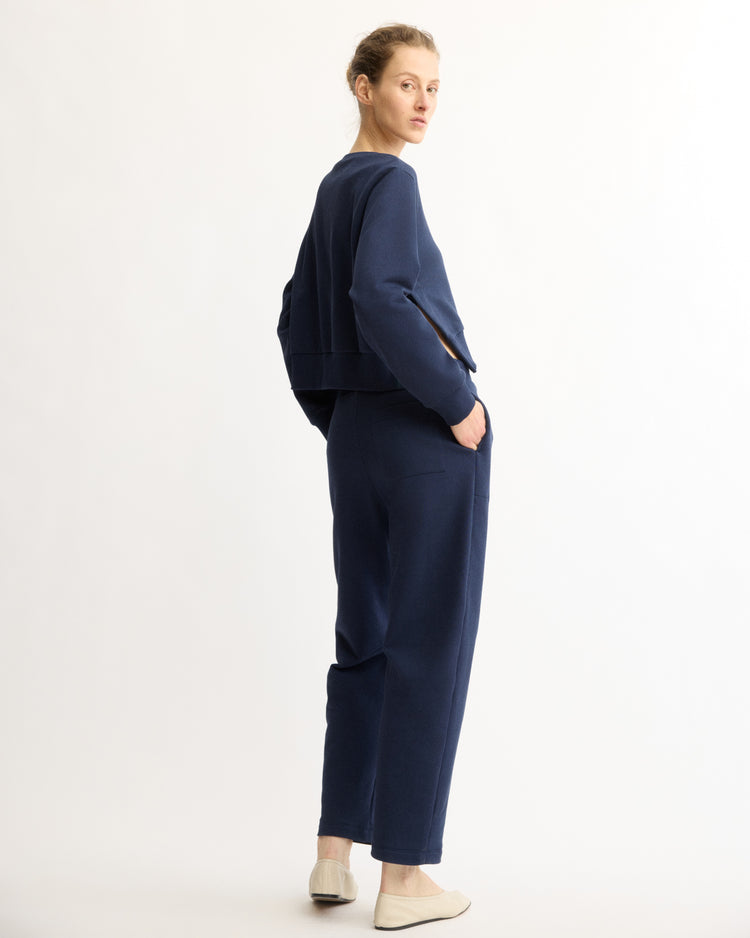 A woman stands sideways against a white background, looking over her shoulder. She wears Arlo Mott's Baltic Barrel Leg Sweatpants in Navy with a matching navy Italian terry top and light slip-on shoes.