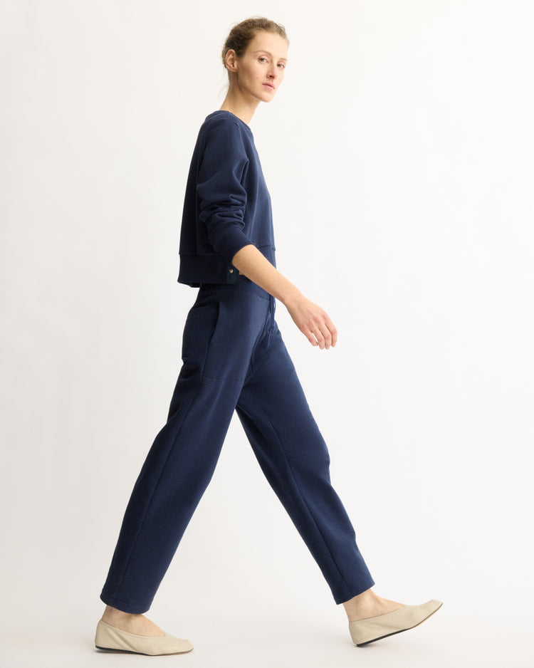 A person in Arlo Mott Baltic Barrel Leg Sweatpants in Navy walks right against a plain white background, paired with a matching navy long-sleeve top and light beige flat shoes.
