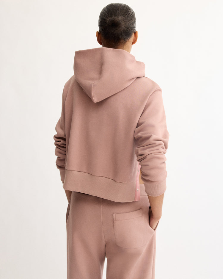 A person with a bun wears the Arlo Mott Morris Hoodie With Snaps in Dusty Rose, paired with matching pants, standing hands-in-pockets and facing away from the camera against a plain white background.