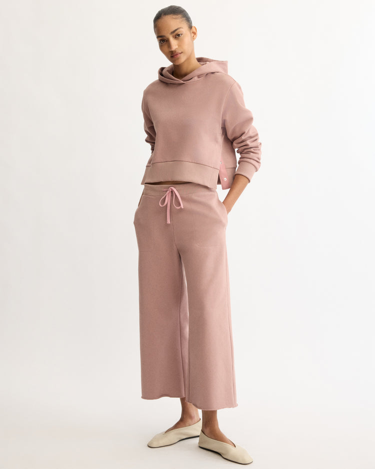 A woman stands against a plain backdrop wearing Arlo Mott's Kane Cropped Wide Sweatpants in Dusty Rose and a matching hooded sweatshirt, both made from Italian terry cloth. Her hands are in her pockets, and she wears cream-colored slippers.