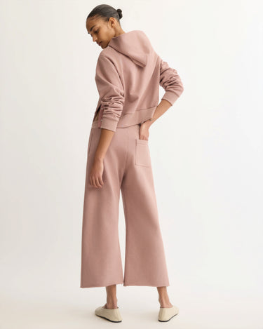A person with their hair in a neat bun wears Arlo Mott's Kane Cropped Wide Sweatpants in Dusty Rose—polished, patch-pocketed, New York-made—paired with a matching hoodie and light slip-on shoes, facing away from the camera.