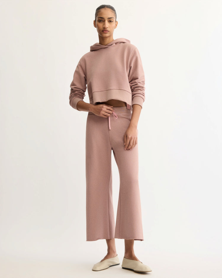 A woman stands against a light background wearing Arlo Mott’s Kane Cropped Wide Sweatpants in Dusty Rose with a matching dusty pink hoodie and cream slip-on shoes.