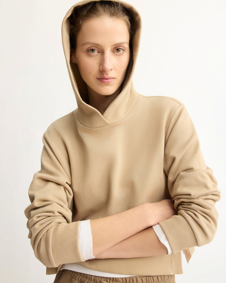 A person with light skin and pulled-back hair wears the Arlo Mott Morris Hoodie With Snaps in Khaki, styled over a white top. They stand with arms crossed, looking at the camera against a plain light background.