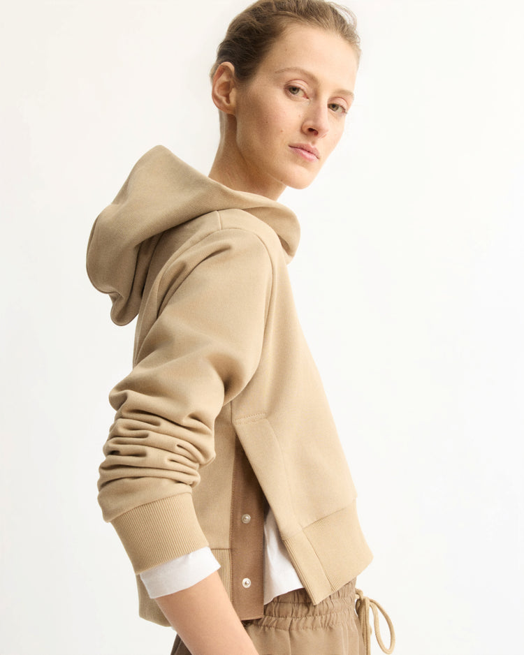 A person stands sideways, looking at the camera in the Arlo Mott Morris Hoodie With Snaps in Khaki and matching joggers. The boxy cropped hoodie features snap details at the side. The background is plain white.