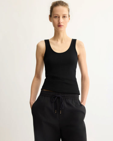 A person with hands in pockets and a neutral expression wears the Arlo Mott Nevins Tank Top in Black against a light background, highlighting its slim silhouette.