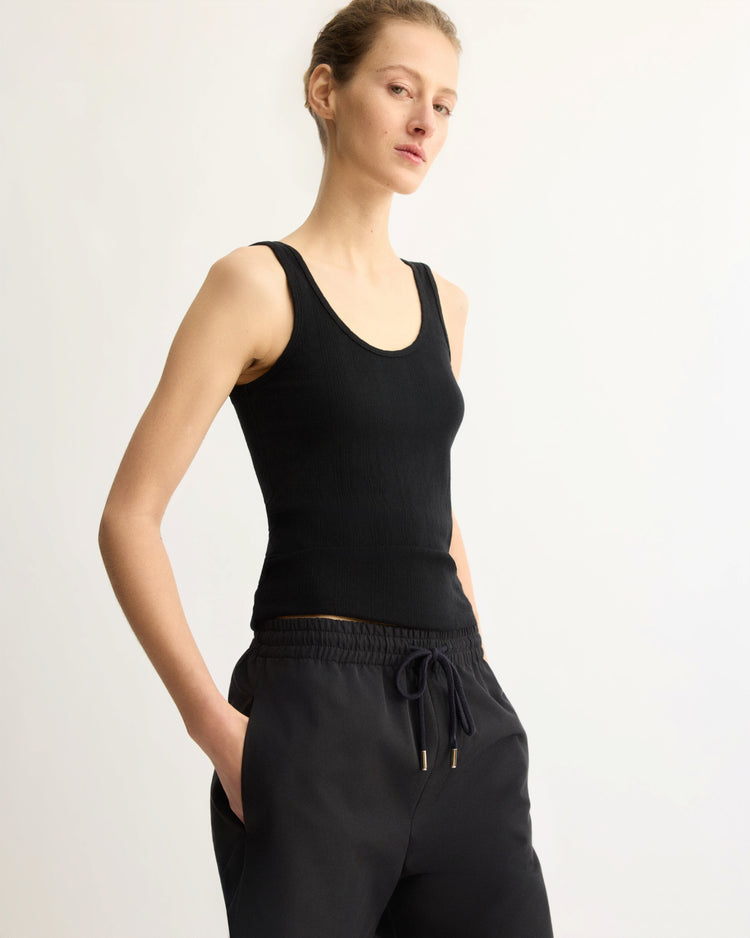 Wearing the Arlo Mott Nevins Tank Top in Black and black drawstring pants, a person with hands in pockets showcases a slim silhouette against a plain light background.
