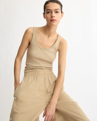 A woman wears the Arlo Mott Nevins Tank Top in khaki with loose beige pants, standing against a white background with one hand in her pocket, highlighting a slim silhouette.