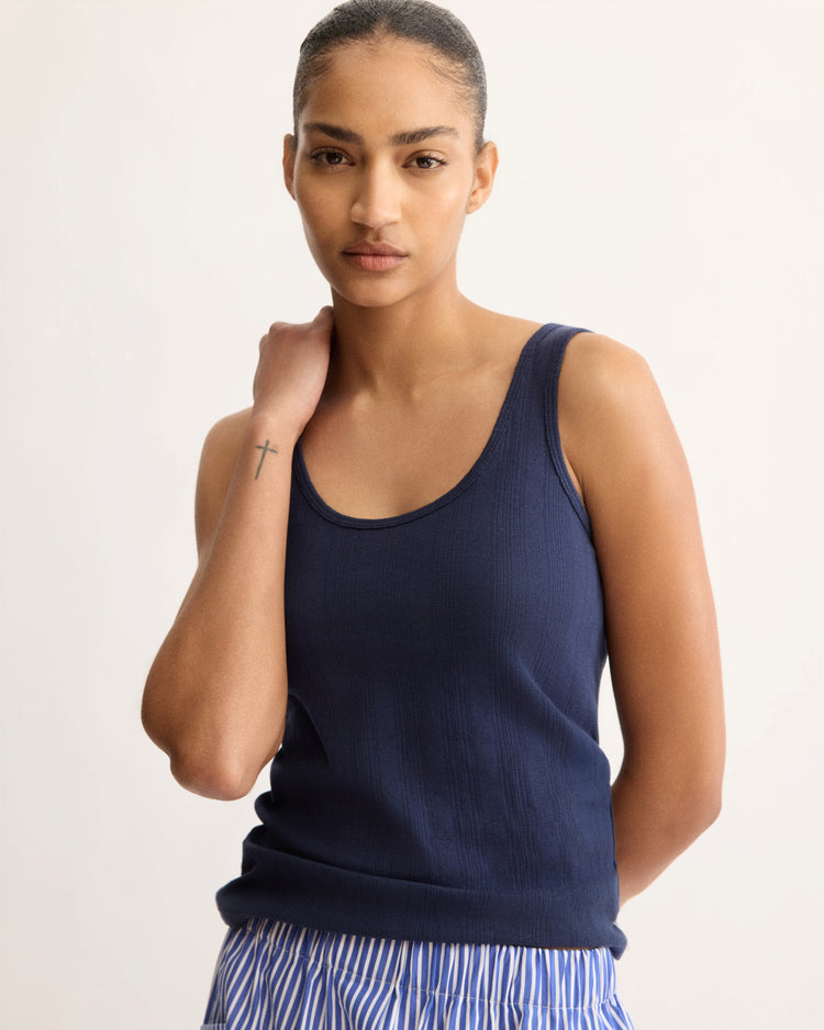 A woman wears the Arlo Mott Nevins Tank Top in Navy with blue-and-white striped pants, standing against a plain light background. She gazes confidently at the camera, hand on her neck, showcasing a slim silhouette and a cross tattoo on her left wrist.