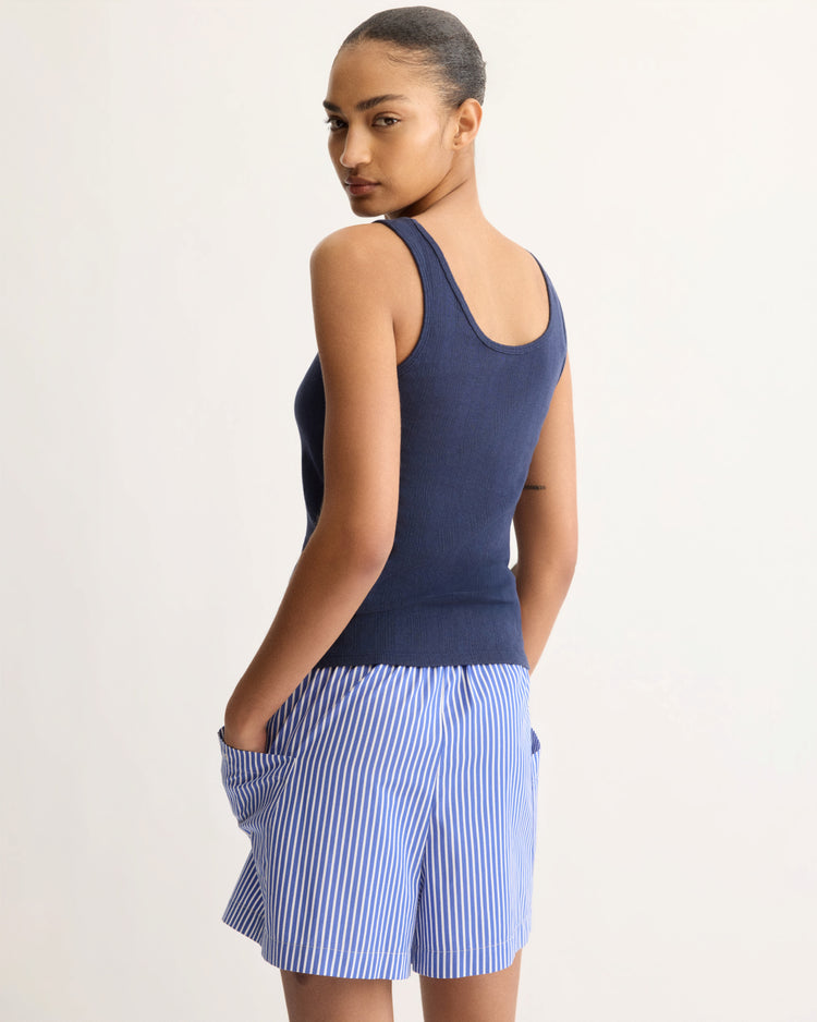 A person stands facing away, looking over their shoulder. They wear the Nevins Tank Top in Navy by Arlo Mott and blue-and-white striped slim shorts, hands in pockets, against a plain light background.