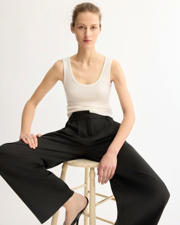 A person with light skin and tied-back hair sits on a stool against a plain white background, wearing the Arlo Mott Nevins Tank Top in White and wide-legged black pants, looking directly at the camera with a neutral expression.