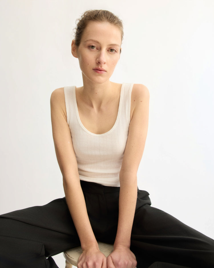 A woman with light skin and tied-back hair sits on a stool against a white backdrop, wearing the Nevins Tank Top in White by Arlo Mott and black pants, gazing calmly at the camera with her hands resting between her knees.