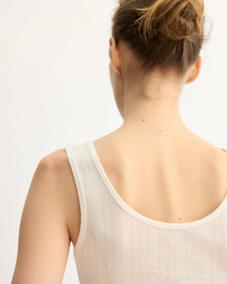 A woman with light skin and brown hair in a bun is seen from behind wearing the Arlo Mott Nevins Tank Top in White, a sleeveless piece that flatters her slim silhouette against a plain light background.