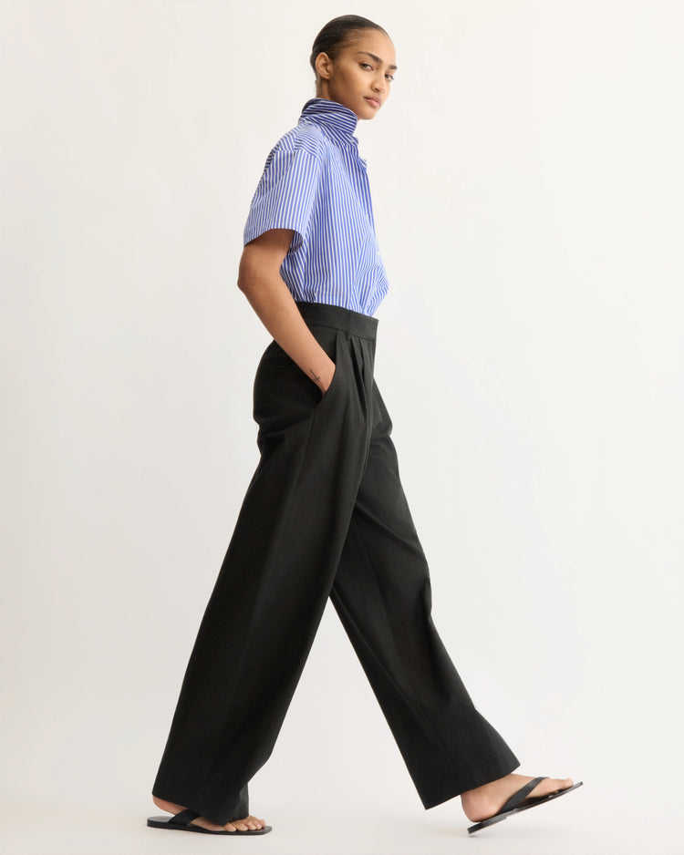 A person wearing a blue and white striped short-sleeve shirt, Arlo Mott Horatio Pleated Barrel Trouser in Black, and black sandals walks confidently against a plain, light background.