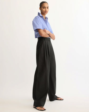 A person stands confidently in a studio, arms folded, wearing a blue and white striped short-sleeve shirt and the Arlo Mott Horatio Pleated Barrel Trouser in Black, paired with black sandals as they look toward the camera.