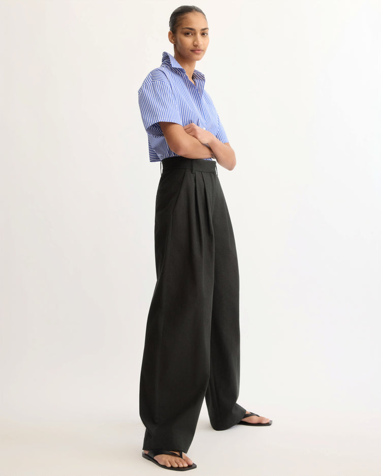 A person stands confidently in a studio, arms folded, wearing a blue and white striped short-sleeve shirt and the Arlo Mott Horatio Pleated Barrel Trouser in Black, paired with black sandals as they look toward the camera.