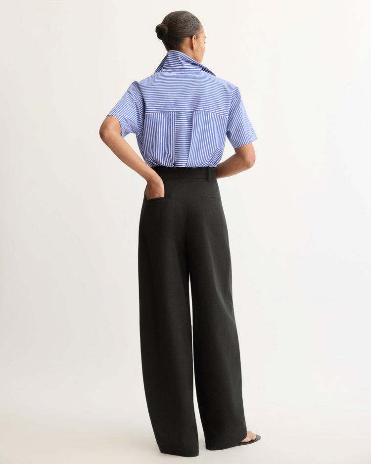 A person stands with their back to the camera, wearing Arlo Mott's Horatio Pleated Barrel Trouser in Black, high-rise with a tapered leg in Japanese twill, paired with a blue striped short-sleeve shirt against a light background.