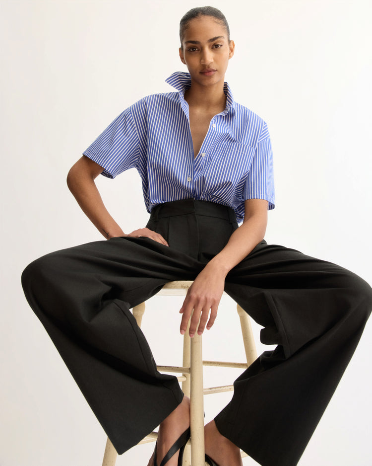 A woman sits on a stool against a plain background, confidently wearing the Arlo Mott Clark Split Back Top in Blue Stripe paired with high-waisted wide-leg black pants.