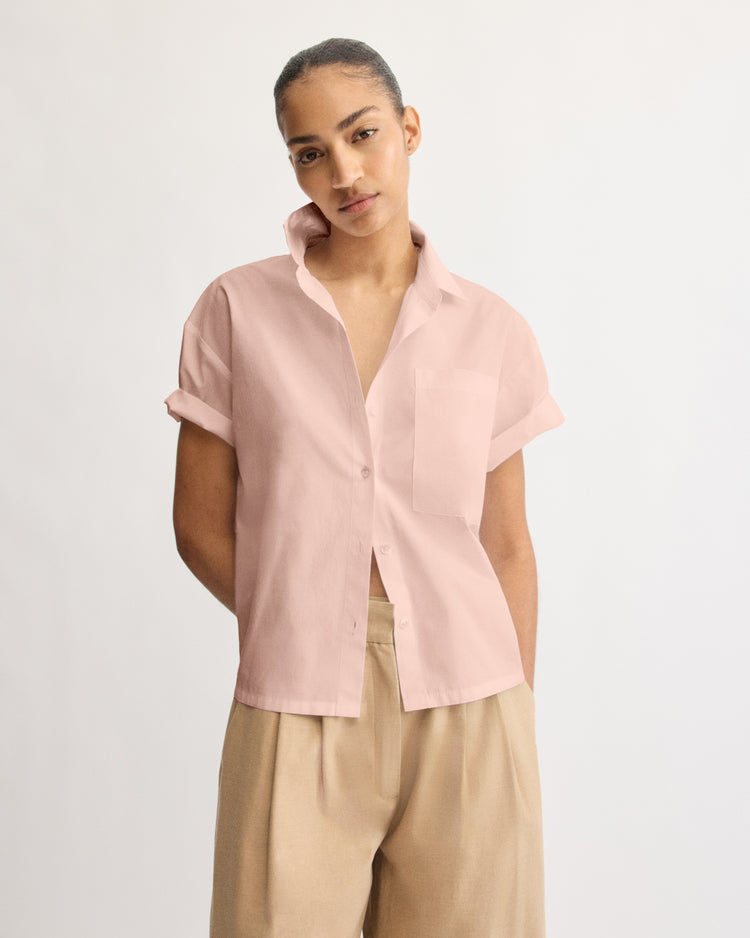 A woman with her hair pulled back and a neutral expression wears the Arlo Mott Clark Split Back Top in Dusty Rose, featuring short sleeves and a spread collar, paired with beige high-waisted pants against a plain background.