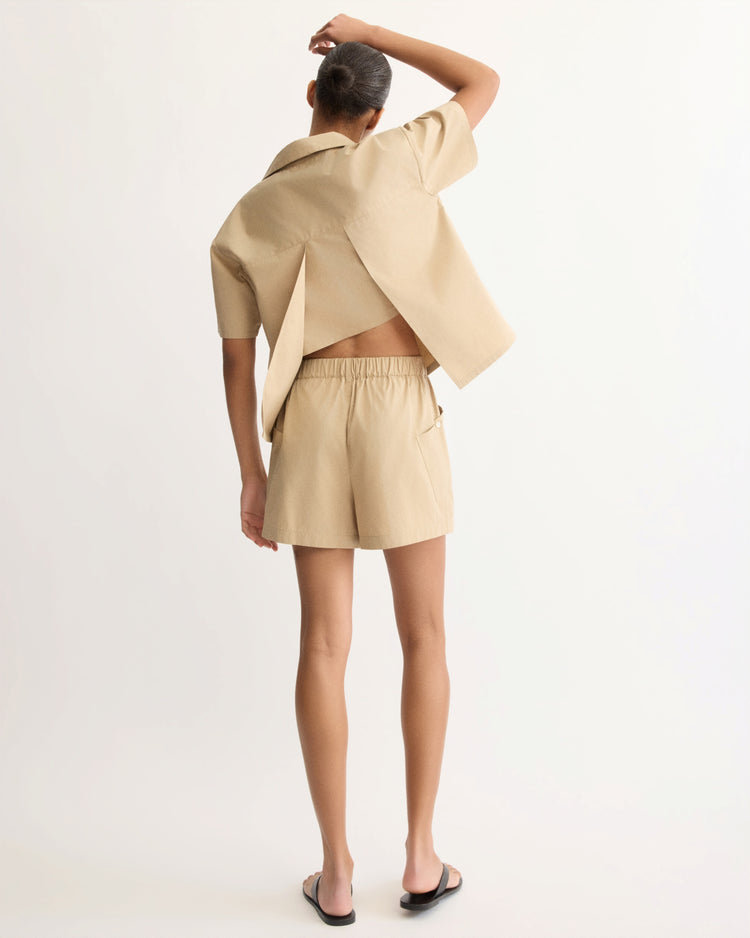 A person faces away, wearing the Arlo Mott Clark Split Back Top in Khaki with matching shorts and black slide sandals, one hand on their head, set against a plain white background.
