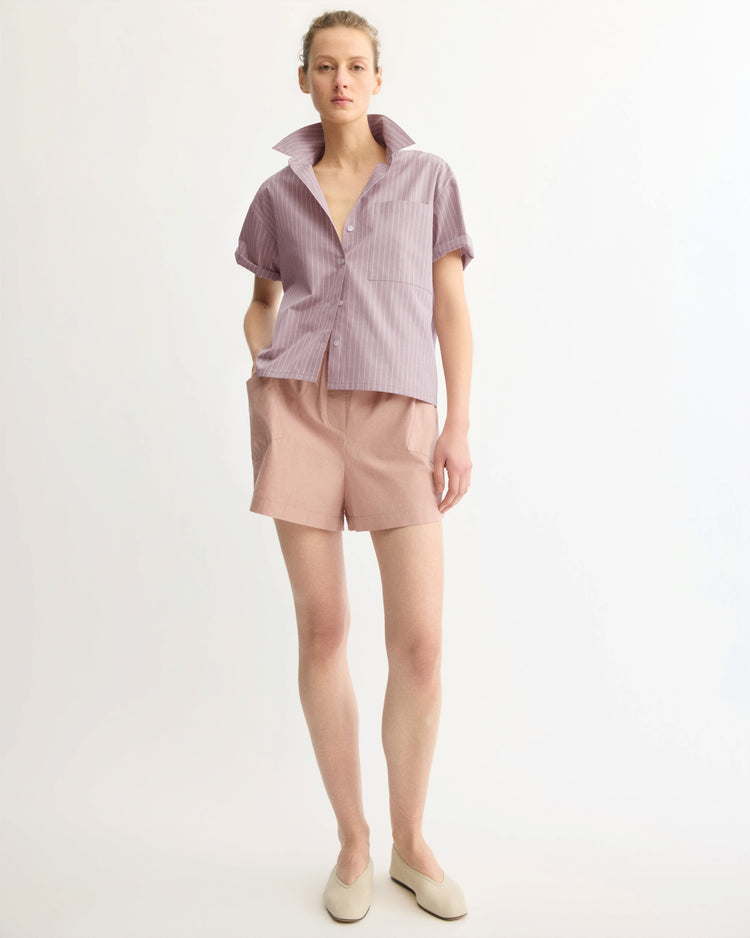 A person stands against a white background, wearing the Arlo Mott Clark Split Back Top in Rose Stripe with light pink shorts and cream slip-on shoes.