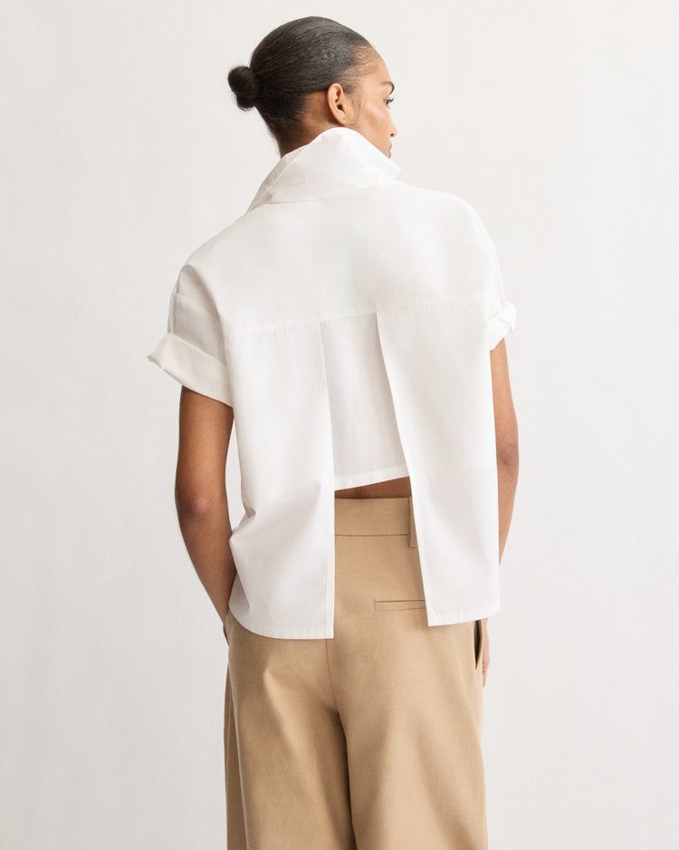 A person with short hair in a bun is seen from the back, wearing the Arlo Mott Clark Split Back Top in White, featuring wide back pleats and Mother of Pearl buttons, paired with tan high-waisted trousers. The background is plain and light-colored.