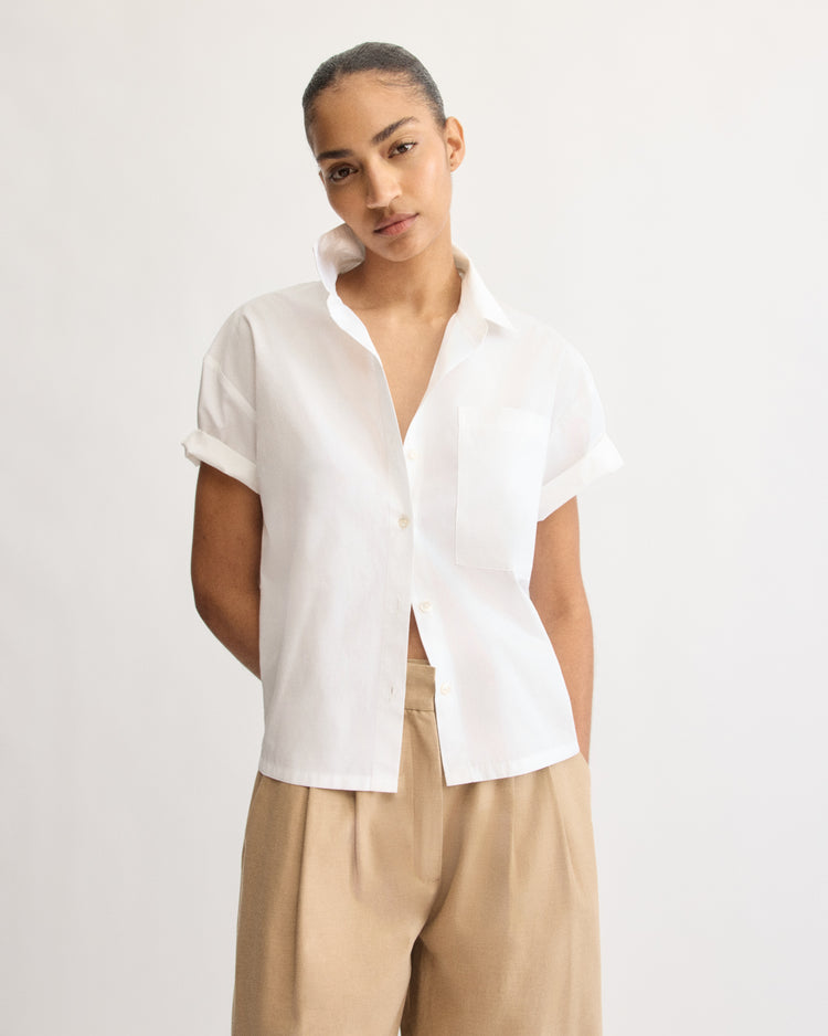 A person stands against a plain background, wearing the Arlo Mott Clark Split Back Top in White and tan wide-leg pants. Their hair is pulled back as they look calmly at the camera.