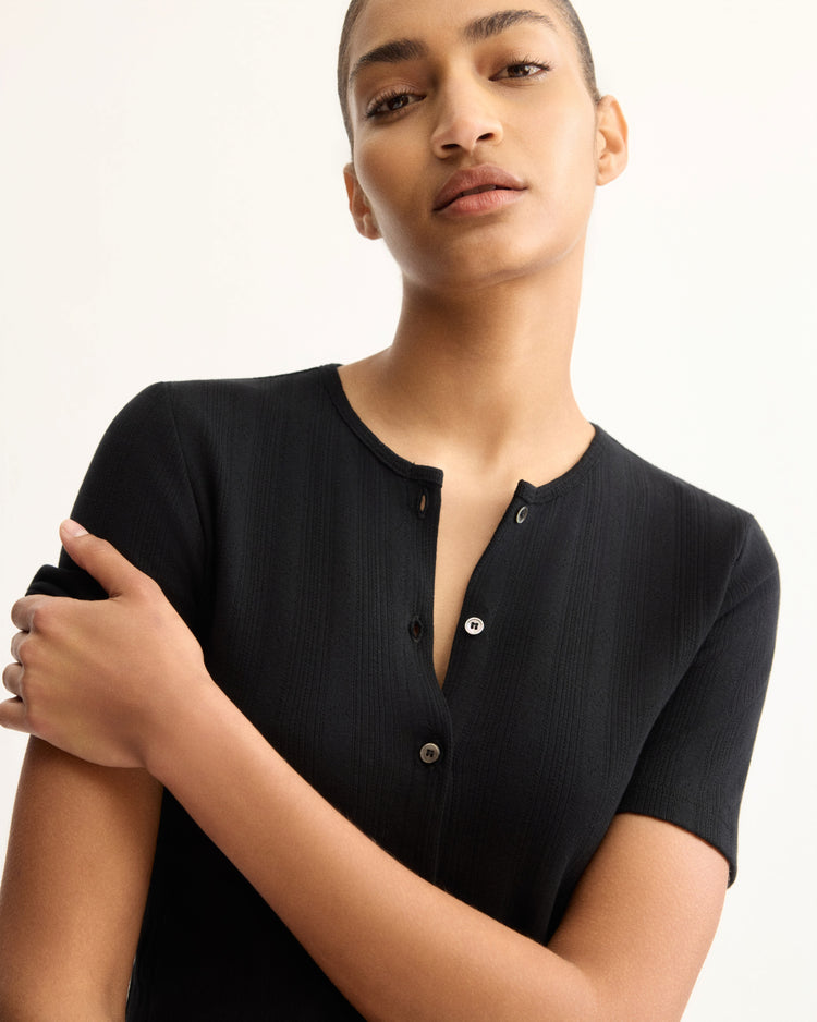 A person wearing the Marion Short Sleeve Cardigan in Black by Arlo Mott stands against a plain light background, gazing confidently at the camera with one arm crossed over their body.