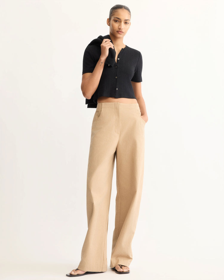 A woman stands against a plain background wearing a black cropped cardigan, black sandals, and Arlo Mott's Leroy Flat Front Barrel Pant in Khaki. She holds a black sweater over her shoulder and has a neutral expression.
