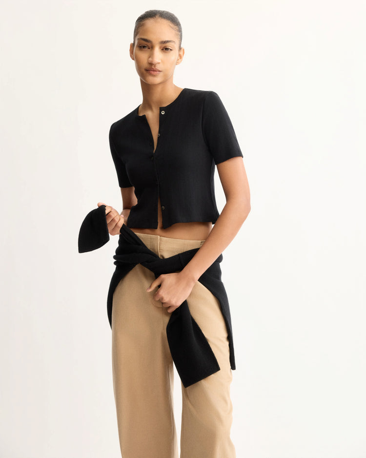 A woman stands against a plain background wearing the Arlo Mott Marion Short Sleeve Cardigan in Black, beige high-waisted pants, with a black sweater tied at her waist. She holds a black beret and looks confidently at the camera.