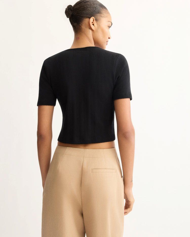 A woman faces away, wearing the Arlo Mott Marion Short Sleeve Cardigan in Black with mother of pearl buttons and high-waisted beige trousers, her hair in a neat bun against a plain light background.