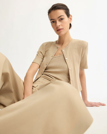 A woman with dark hair sits against a white background, wearing the Arlo Mott Marion Short Sleeve Cardigan in Khaki over a matching top and wide-legged pants, looking confidently at the camera.