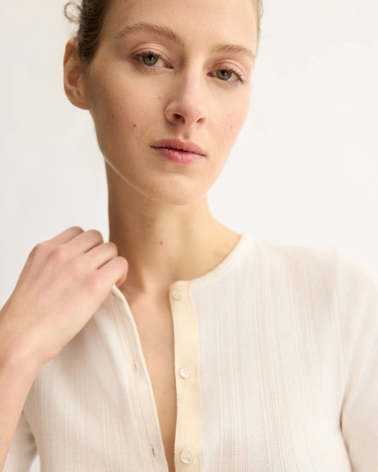 A woman with light skin and light brown hair pulled back wears the Arlo Mott Marion Short Sleeve Cardigan in White. The cream cardigan features mother of pearl buttons. She looks calmly at the camera, her left hand touching her collar against a light background.