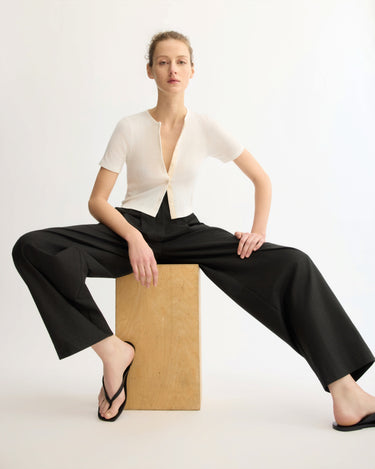 A woman sits confidently on a wooden block in front of a plain background, wearing the Arlo Mott Marion Short Sleeve Cardigan in White, black wide-leg pants, and black flip-flops, with one foot on the ground and one on the block.