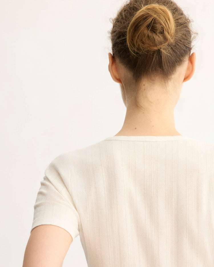 A person with light brown hair in a messy bun is shown from behind, wearing the Arlo Mott Marion Short Sleeve Cardigan in White with delicate mother of pearl buttons, set against a plain light background.