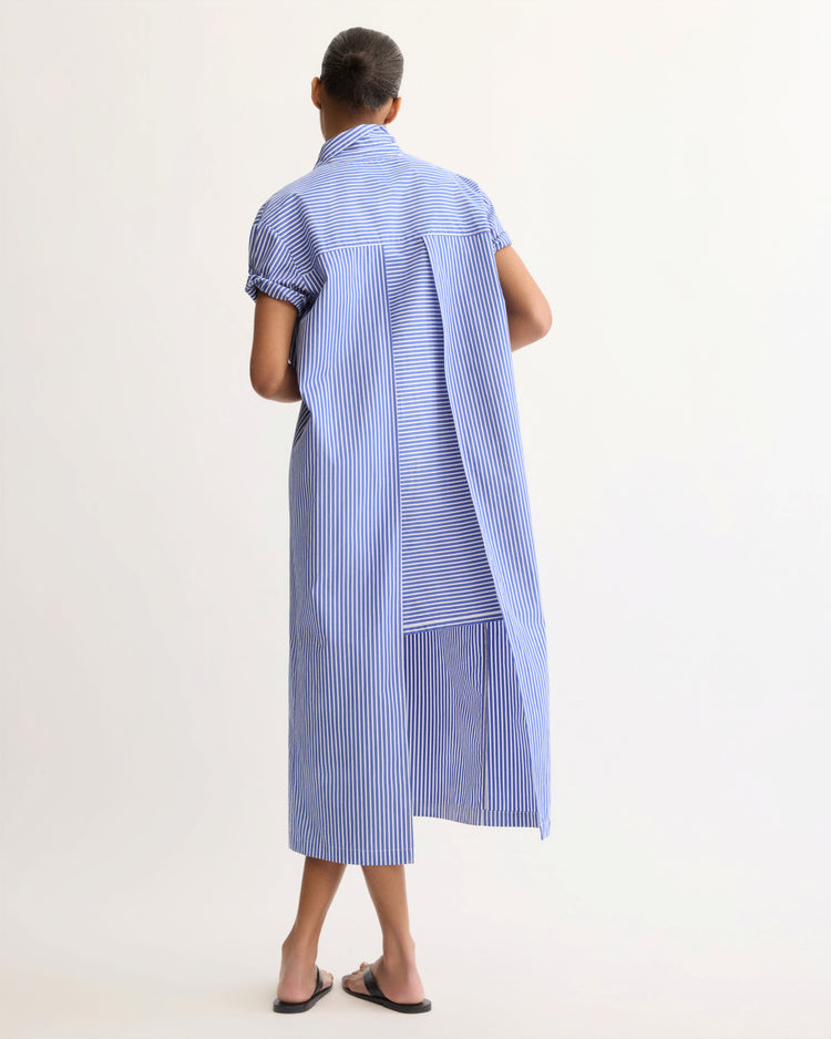 A person with short hair wears the Emerson Split Back Dress in Blue Stripe by Arlo Mott, featuring vertical stripes, pleats, a spread collar, and split back, paired with black flat sandals against a plain light background.