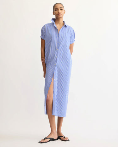 A person wears the Arlo Mott Emerson Split Back Dress in Blue Stripe—featuring short sleeves and a front slit—and pairs it with black sandals, standing with one leg forward.