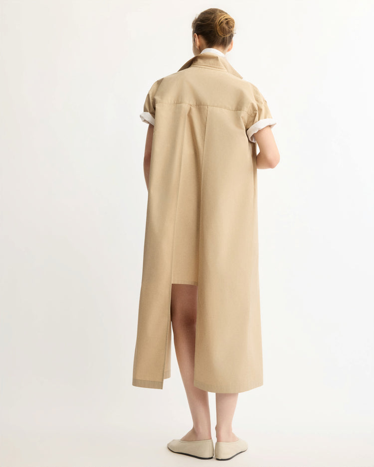 A woman faces away in the Arlo Mott Emerson Split Back Dress in Khaki, featuring short sleeves, rolled cuffs, side seam pockets, and a back slit, paired with light flat shoes against a plain white background.