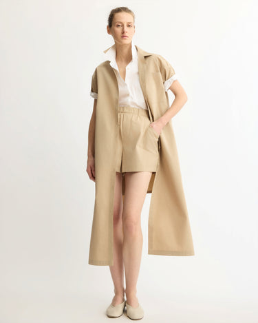 A woman poses against a plain background wearing the Arlo Mott Emerson Split Back Dress in Khaki, styled with beige flats and one hand in her pocket.
