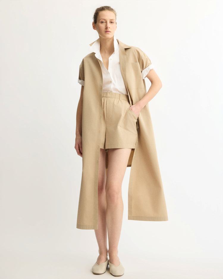 A woman poses against a plain background wearing the Arlo Mott Emerson Split Back Dress in Khaki, styled with beige flats and one hand in her pocket.