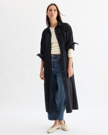 A woman poses against a white background in the Arlo Mott Lafayette Side Button Maxi Dress in Black, layered over a beige top and blue high-waisted jeans with cream shoes. Her hair is straight and worn loose.