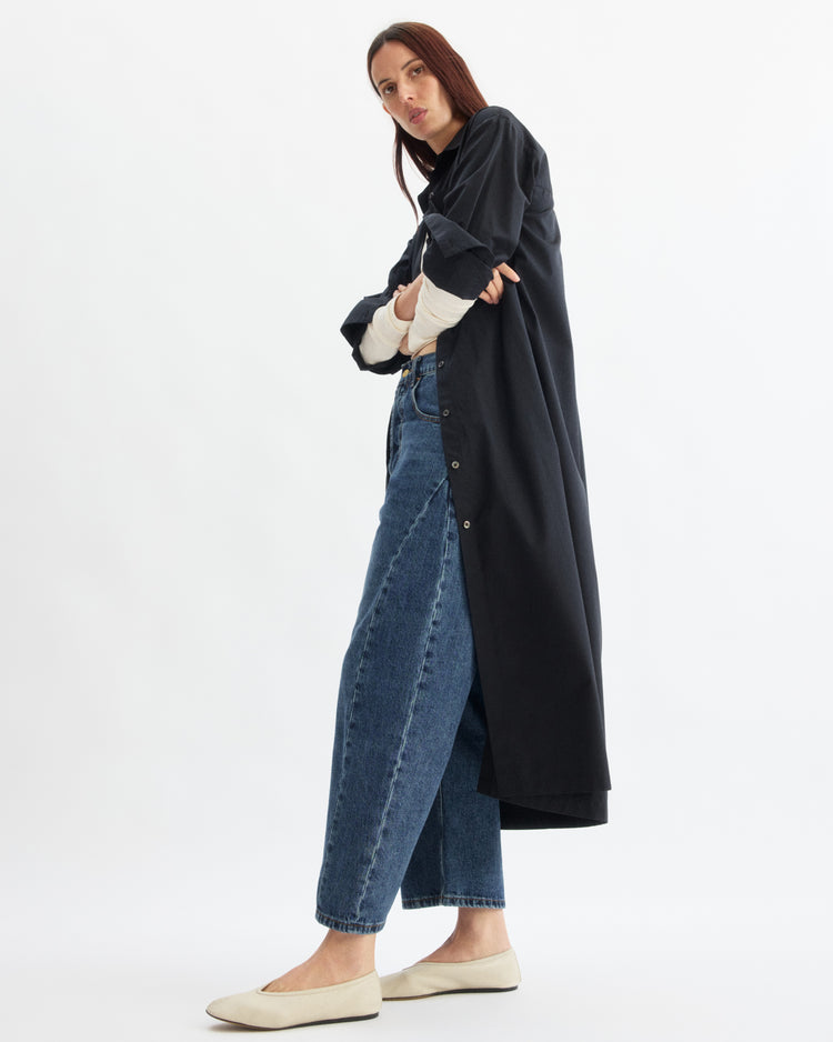 A woman stands against a white background, wearing the Arlo Mott Lafayette Side Button Maxi Dress in Black over a light long-sleeve top, loose blue jeans, and white flats. She stands with one arm folded, looking slightly down.