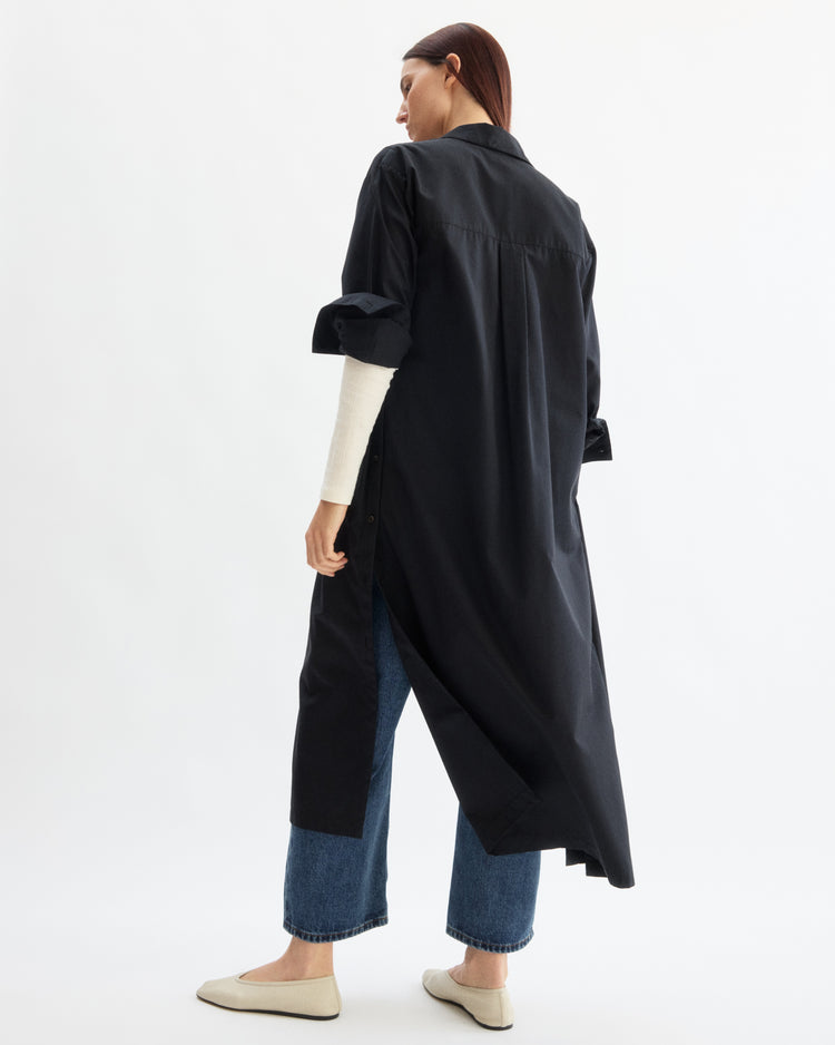A person stands facing away, wearing the Arlo Mott Lafayette Side Button Maxi Dress in Black with rolled-up sleeves over a white top, blue jeans, and beige flats against a plain white background.