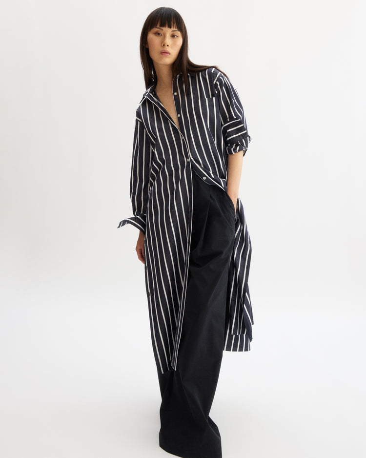 A woman stands against a white background wearing the Arlo Mott Lafayette Side Button Maxi Dress in Navy Stripe. The shirt sleeves are rolled up, and she poses with one hand in her pocket.
